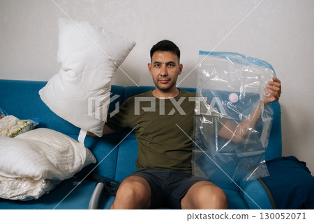 Portrait of young man packing white pillows and bedding into vacuum storage bags sitting on blue sofa in living room, getting ready for moving or tidying up. Concept of space saving storage. 130052071