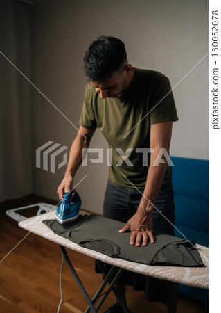 Vertical shot of focused man ironing clothes on ironing board in cozy living room, managing household chores and ensuring garments neat and wrinkle-free. Concept of domestic work, housekeeping Vertical shot of focused man ironing clothes on ironing board in cozy living room, managing household chores and ensuring garments neat and wrinkle-free. Concept of domestic work, housekeeping 130052078