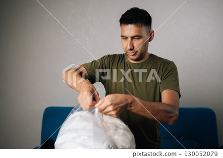 Focused young man closing large transparent vacuum storage bag filled with white bedding, using double-zip closure to create airtight seal for optimal space efficiency and protection. Focused young man closing large transparent vacuum storage bag filled with white bedding, using double-zip closure to create airtight seal for optimal space efficiency and protection. 130052079