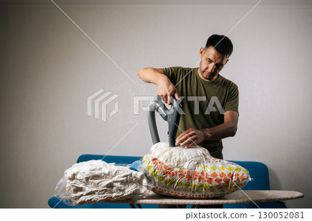 Portrait of man vacuuming compressed storage bag with bedding on ironing board, demonstrating efficient home organization and space-saving technique. Concept of space saving and careful storage 130052081