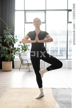 Woman in black sportswear practicing tree pose during yoga session in a bright room with large windows. Woman in black sportswear practicing tree pose during yoga session in a bright room with large windows. 130052906