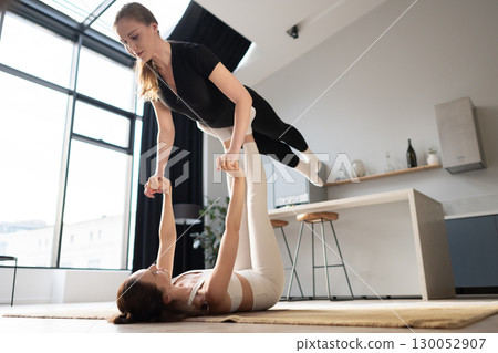 Two women practicing acro yoga at home, one lifting the other in a balance pose with teamwork and strength. 130052907