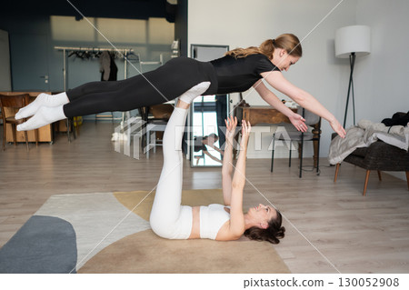 Sporty women doing acro yoga balance training at home, performing stretching and strength exercise with concentration, trust, and teamwork. Sporty women doing acro yoga balance training at home, performing stretching and strength exercise with concentration, trust, and teamwork. 130052908