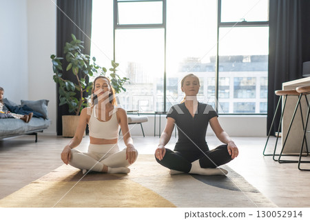 Two women practicing yoga and meditation at home, sitting in lotus pose, calm breathing and relaxation exercise in natural light. 130052914