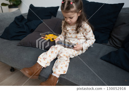 Girl sits alone on sofa at home, eating fruit and watching smartphone Girl sits alone on sofa at home, eating fruit and watching smartphone 130052916