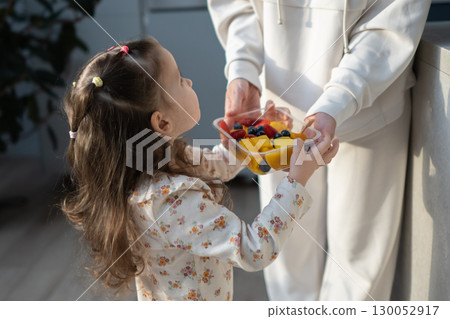 Mother and child in home kitchen, girl takes fruit from container 130052917