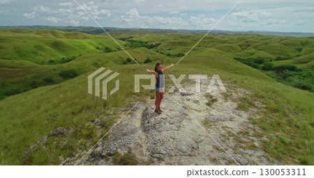 Young woman standing on top of a hill with open arms, embracing the beauty of rolling green hills and lush vegetation of Sumba Island under a cloudy sky Young woman standing on top of a hill with open arms, embracing the beauty of rolling green hills and lush vegetation of Sumba Island under a cloudy sky 130053311