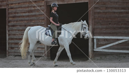 Female equestrian wearing a helmet and casual attire riding a beautiful white horse while leaving the stable at a ranch, enjoying the freedom of the outdoors and equine companionship Female equestrian wearing a helmet and casual attire riding a beautiful white horse while leaving the stable at a ranch, enjoying the freedom of the outdoors and equine companionship 130053313