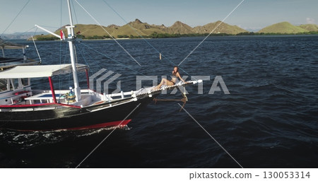 Aerial drone footage of a young tourist relaxing and sunbathing on the bowsprit of a yacht sailing in the beautiful Komodo National Park, Indonesia 130053314