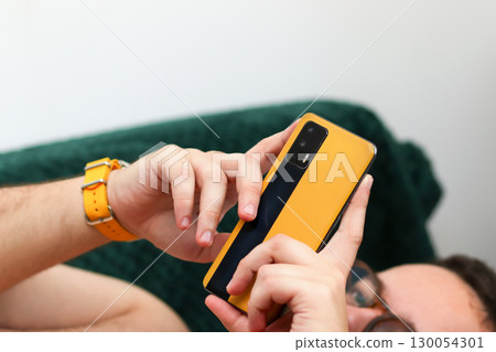 a man holding a yellow smartphone while lies on a couch at home. man using smartphone home couch. casual person with phone relaxation a man holding a yellow smartphone while lies on a couch at home. man using smartphone home couch. casual person with phone relaxation 130054301