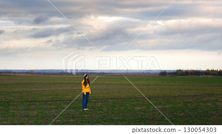a woman standing in a green meadow. a beautiful autumn day and a vibrant yellow raincoat. woman yellow raincoat field autumn. solitude in nature fall fashion 130054303