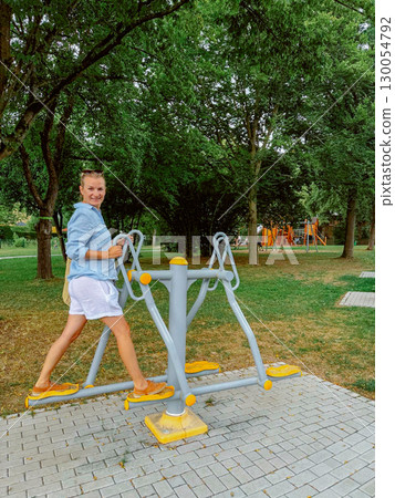 Woman using outdoor fitness equipment in a park setting. Her expression is cheerful and focused on her workout. Surrounding greenery provides a pleasant environment for exercise. Woman using outdoor fitness equipment in a park setting. Her expression is cheerful and focused on her workout. Surrounding greenery provides a pleasant environment for exercise. 130054792