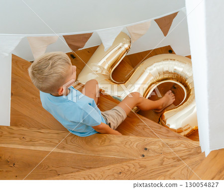 A young boy wearing a blue shirt and beige shorts sits on wooden stairs, holding a large gold-colored balloon in the shape of a number 6. Party decorations hang above. 130054827