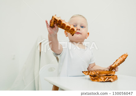 A happy baby joyfully enjoys a delicious waffle at the table, surrounded by a cheerful atmosphere that radiates happiness, showcasing precious moments of childhood and family togetherness 130056047