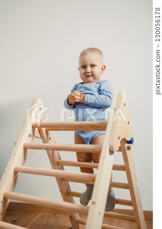 An absolutely adorable baby is happily standing upright on a colorful wooden climbing frame situated in a modern playroom, showcasing the carefree spirit and joy of early childhood and playtime 130056178