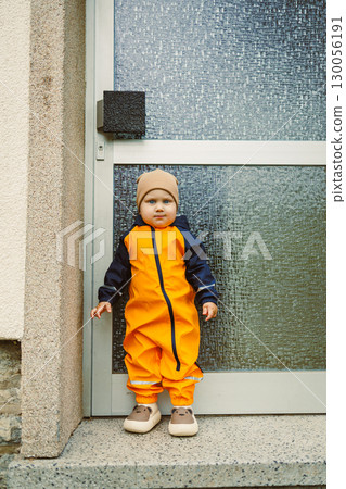 An Adorable Child Dressed in a Bright Orange Rain Suit is Standing Just Beside a Large Glass Door in an Urban Setting, Showcasing Their Innocent and Playful Nature Amidst Vibrant Surroundings An Adorable Child Dressed in a Bright Orange Rain Suit is Standing Just Beside a Large Glass Door in an Urban Setting, Showcasing Their Innocent and Playful Nature Amidst Vibrant Surroundings 130056191