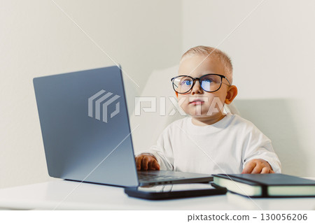 A 2-year-old boy dressed as a businessman in glasses sits at a table and works on a laptop. A toddler in a white T-shirt sits and watches a video on a laptop. A child works on a laptop. 130056206