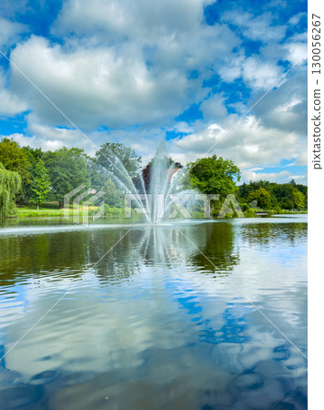 This serene park features a charming fountain amidst lush greenery, reflected in a tranquil pond under a blue sky. Its an ideal spot for relaxation, photography, and enjoying nature in summer This serene park features a charming fountain amidst lush greenery, reflected in a tranquil pond under a blue sky. Its an ideal spot for relaxation, photography, and enjoying nature in summer 130056267