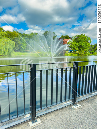 A beautifully tranquil park landscape featuring stunning fountain reflections on the serene still water, elegantly surrounded by lush, vibrant trees and a breathtakingly scenic sky above 130056268