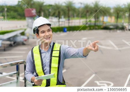 Male engineer in white hardhat standing and holding tablet working aircraft maintenance mechanics moving through hangar. Male engineer in white hardhat standing and holding tablet working aircraft maintenance mechanics moving through hangar. 130056695