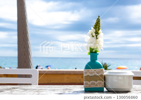 Warehouse on the open air with people. Chairs and tables outside on the summer sun weather. Vase with the flowers on the table for summer cafe with sea view in Spain. Warehouse on the open air with people. Chairs and tables outside on the summer sun weather. Vase with the flowers on the table for summer cafe with sea view in Spain. 130056860