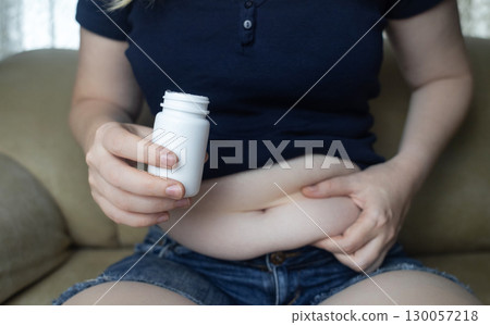 An overweight woman with folds of fat sits on a sofa and holds a jar of diet pills. The image symbolizes the use of diet pills, weight loss pills, and fat burning pills 130057218