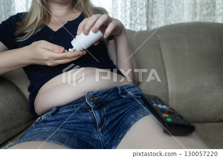 An overweight woman with folds of fat sits on a sofa and holds a jar of diet pills. The image symbolizes the use of diet pills, weight loss pills, and fat burning pills 130057220