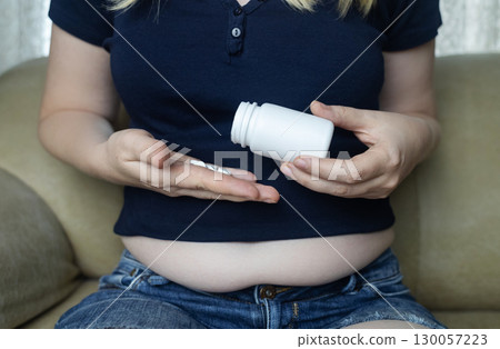 An overweight woman with folds of fat sits on a sofa and holds a jar of diet pills. The image symbolizes the use of diet pills, weight loss pills, and fat burning pills 130057223