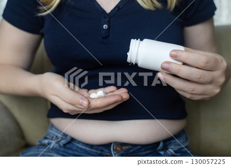 An overweight woman with folds of fat sits on a sofa and holds a jar of diet pills. The image symbolizes the use of diet pills, weight loss pills, and fat burning pills 130057225