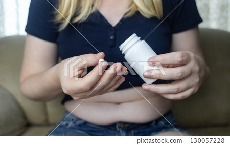 An overweight woman with folds of fat sits on a sofa and holds a jar of diet pills. The image symbolizes the use of diet pills, weight loss pills, and fat burning pills An overweight woman with folds of fat sits on a sofa and holds a jar of diet pills. The image symbolizes the use of diet pills, weight loss pills, and fat burning pills 130057228