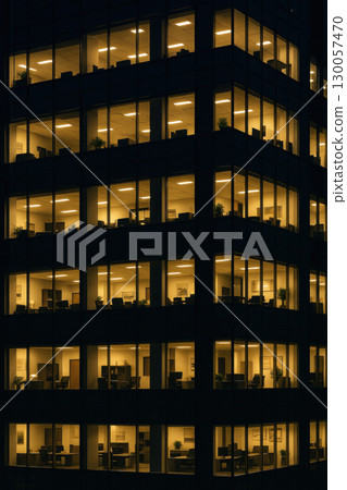 Office building at night with illuminated windows showing empty workspaces and desks in modern corporate environment 130057470
