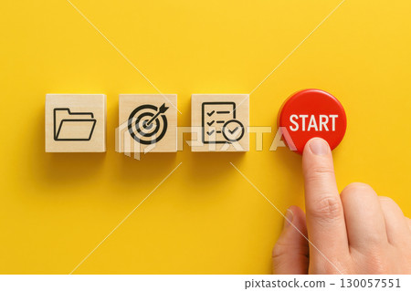 Finger pressing red start button beside wooden blocks with folder target and checklist icons project initiation concept on yellow background 130057551