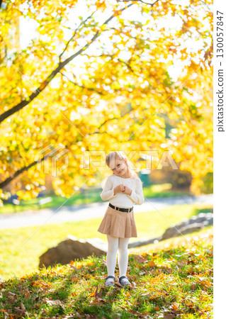 Little girl in autumn park surrounded by colorful foliage, seasonal childhood walk in nature with warm clothing and fallen leaves on the ground 130057847