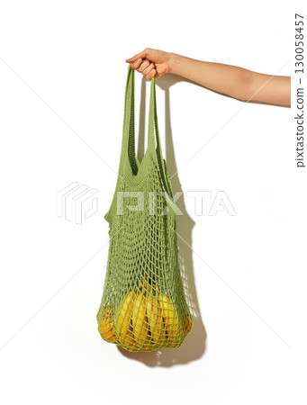 Woman hand holding green lime string shopping bag with vegetables and fruits in front of white wall. Eco lifestyle Woman hand holding green lime string shopping bag with vegetables and fruits in front of white wall. Eco lifestyle 130058457
