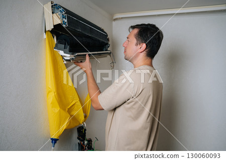 Side view of technician disassembling and repairing wall-mounted air conditioner unit, using drainage bag to collect water while working on electrical components. Concept of air condition hygiene. 130060093
