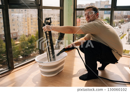 Portrait of professional cleaning service worker wearing safety glasses using steam cleaner to clean air conditioner filter in bucket of water, performing maintenance on HVAC system. 130060102