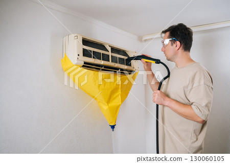 Air conditioning technician in safety glasses cleaning air conditioner unit using steam cleaner. While yellow protective bag collects dirty water. Ensuring thorough and professional cleaning process. 130060105