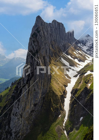 Dramatic mountain peak rises above green valleys and snowy slopes in bright daylight during summer season 130060851