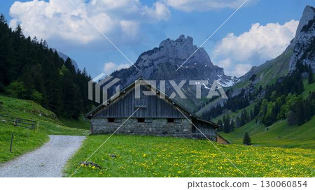 Beautiful mountain landscape with a rustic cabin surrounded by green fields and majestic peaks under a clear sky Beautiful mountain landscape with a rustic cabin surrounded by green fields and majestic peaks under a clear sky 130060854