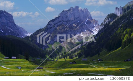Majestic mountains rise above a lush valley with scattered farmhouses in a picturesque landscape under blue skies 130060878
