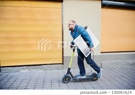 Man rides electric scooter while carrying solar panel. Integration of solar power as sustainable energy source for charging electric scooters, promoting eco-friendly urban transportation. 130061624