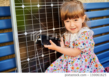 Young girl holds smartphone connected to solar panel for charging. Integration of sustainable renewable energy into daily life, importance of teaching children about eco-friendly technology. Young girl holds smartphone connected to solar panel for charging. Integration of sustainable renewable energy into daily life, importance of teaching children about eco-friendly technology. 130061625