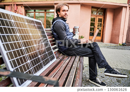 Man sits on wooden bench in urban setting, working on laptop with cup of coffee in hand. Solar panel rests beside, emphasizing sustainable, mobile workspace amidst backdrop of historic architecture. Man sits on wooden bench in urban setting, working on laptop with cup of coffee in hand. Solar panel rests beside, emphasizing sustainable, mobile workspace amidst backdrop of historic architecture. 130061626