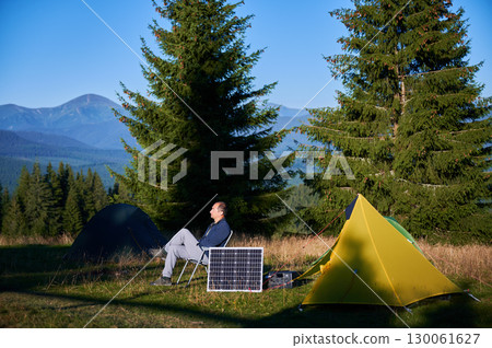 Man using photovoltaic solar panel for charging portable power station outdoor in eco-friendly camping. Male tourist sits relaxed in chair near tents. On background tall pine trees and clear blue sky. Man using photovoltaic solar panel for charging portable power station outdoor in eco-friendly camping. Male tourist sits relaxed in chair near tents. On background tall pine trees and clear blue sky. 130061627