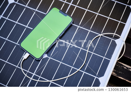 Smartphone with green screen connected to solar panel via charging cable. Image highlights use of solar energy for charging electronic devices, emphasizing renewable and portable power solutions. 130061653