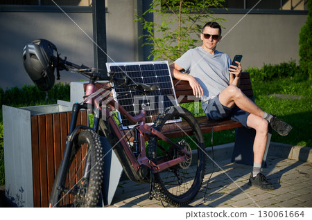 Man cyclist using solar panel for charging electric mountain bike outdoors. Guy in sunglasses sits on bench, holding phone, casually dressed in t-shirt, shorts, enjoying sunny outdoor setting. 130061664