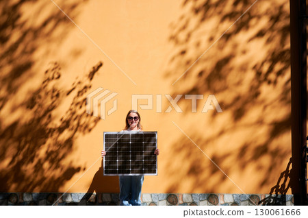 Woman holding solar panel for charging, stands against vibrant orange wall. Smiling girl wears sunglasses, white top, blue jeans. Artistic shadows cast by nearby trees branches on wall. 130061666