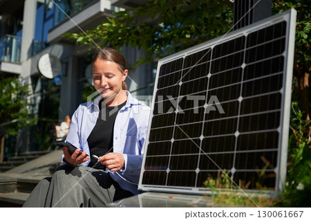 Happy woman connecting smartphone to photovoltaic solar panel. Integration of sustainable renewable energy into everyday life, demonstrating practical use of solar power for charging devices. 130061667