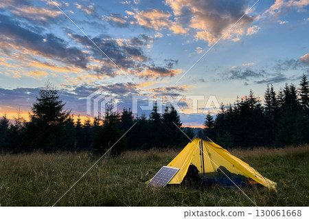 Yellow tent pitched in grassy field with solar panel beside it, capturing last light of setting sun. Backdrop of silhouetted trees and colorful sky emphasizes eco-friendly camping in nature. 130061668
