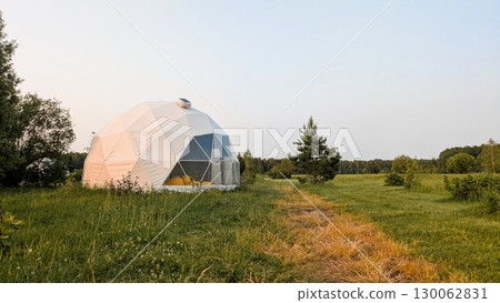 Geodesic dome glamping tent sits in grassy field with path leading to it. Large white tent has clear front panel and forest is visible in background under pale hazy sky 130062831